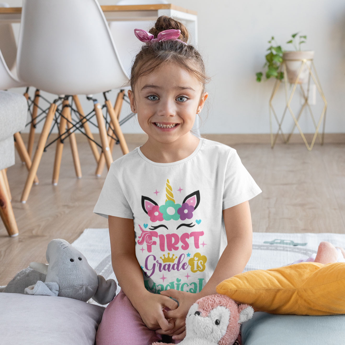 Young girl in a room with a 'First Grade is Magical' shirt, surrounded by stuffed animals.