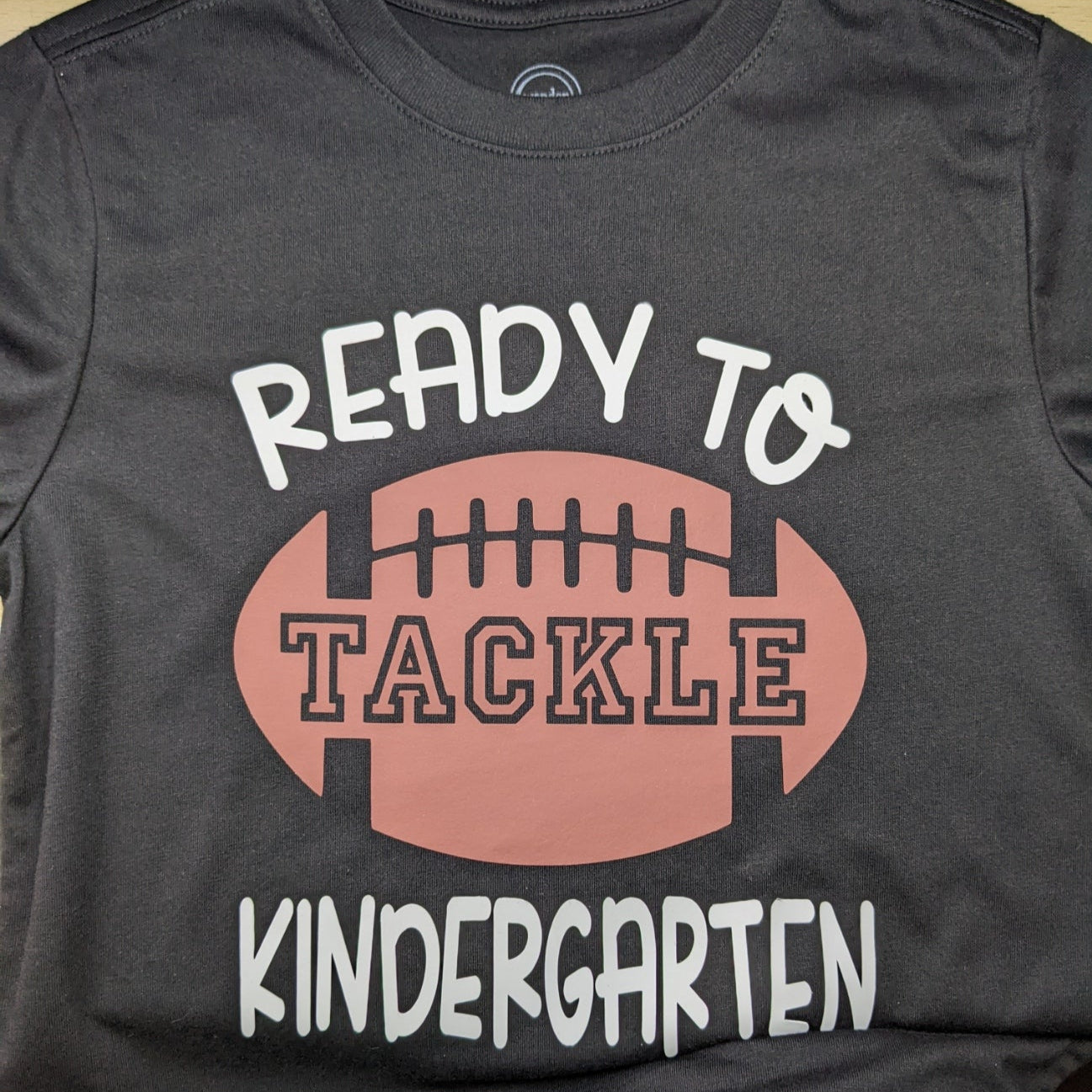 Black t-shirt with 'Ready to Tackle Kindergarten' text and football graphic on a wooden surface